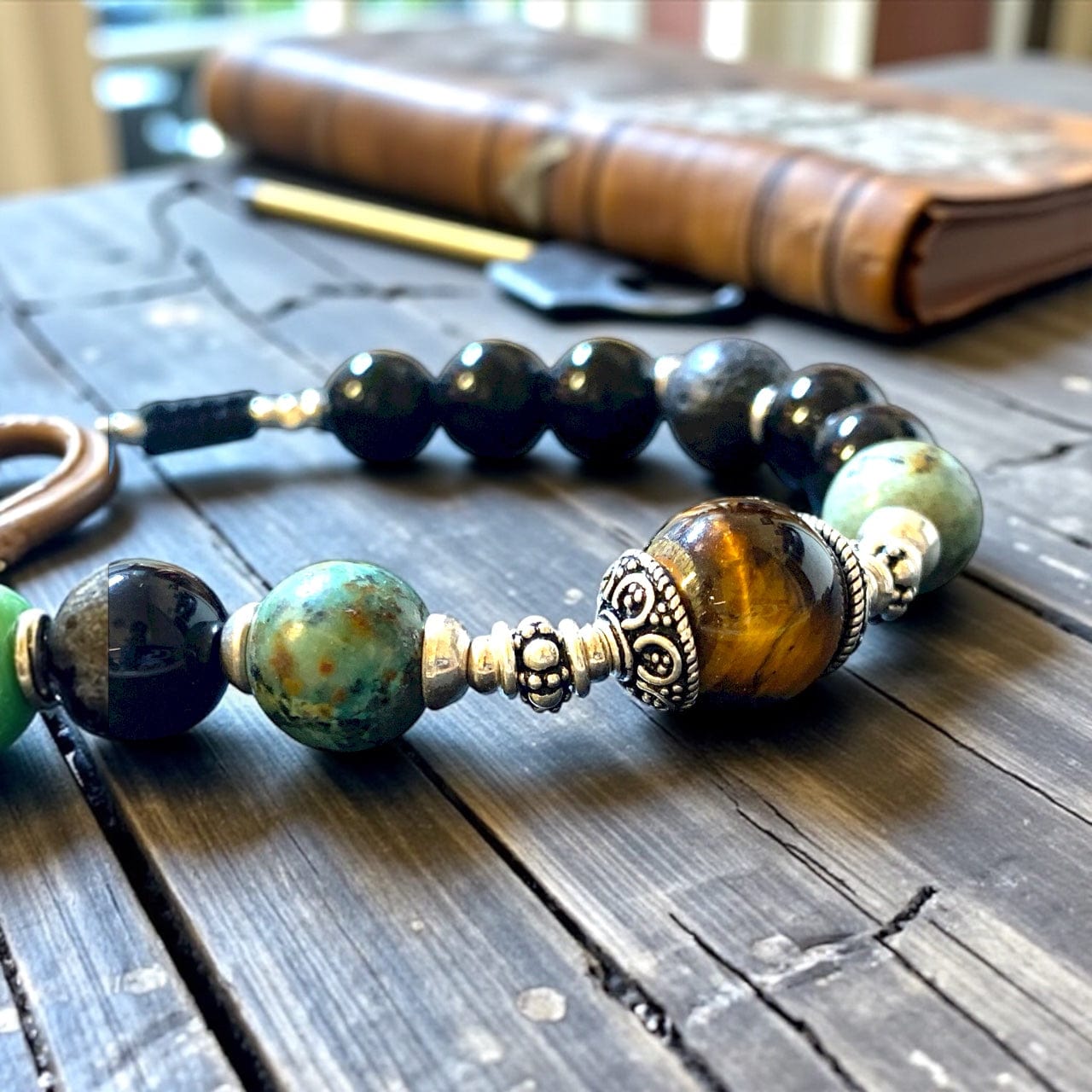 Gros plan bracelet d'un bracelet hommes en pierres naturelles, oeil de tigre, turquoise africaine, onyx et argent 925 posé sur du bois avec un livre en cuir en fond
